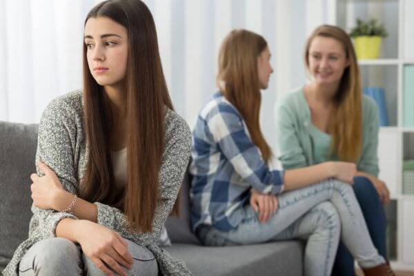 Young woman with a sad expression and crossed arms in the foreground, feeling excluded while two friends converse in the background, symbolizing resentment and the need for emotional healing.