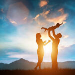 A silhouette of a family enjoying a moment of happiness at sunset. A father holds his young child above his head, while the mother stands nearby, looking at the child with affection. The background features rolling hills, mountains, and an orange-tinged blue sky, symbolizing harmony and joy.