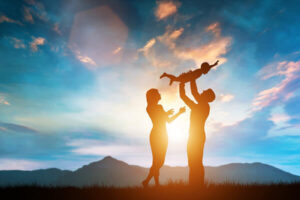 A silhouette of a family enjoying a moment of happiness at sunset. A father holds his young child above his head, while the mother stands nearby, looking at the child with affection. The background features rolling hills, mountains, and an orange-tinged blue sky, symbolizing harmony and joy.