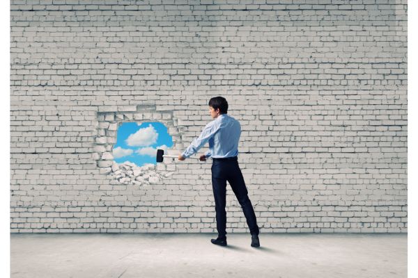 A man in a blue shirt and dress pants uses a sledgehammer to break a white-painted brick wall, revealing a blue sky through the hole, representing the overcoming of barriers in the grieving process.