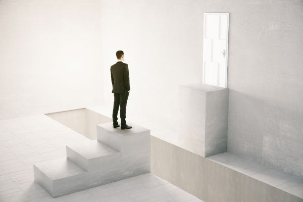 A man in a suit stands on concrete steps, blocked by a light wooden cube, with a closed white door in the background, symbolizing a path obstructed by grief.