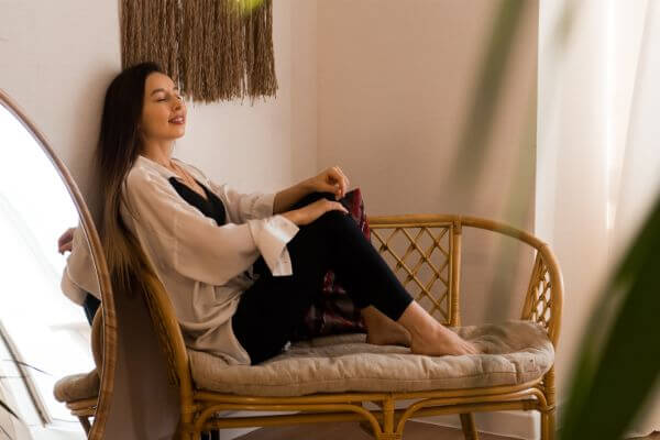 Young brunette woman experiencing well-being, comfortably seated on a bamboo bench with a light cushion, eyes closed and serene expression, in a cozy, naturally lit indoor setting.