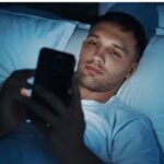 Young man lying in bed in the dark, using his cell phone with the blue light from the screen illuminating his face. The image illustrates bad habits of using smartphones before bed, highlighting the impacts on sleep quality.