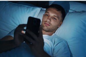 Young man lying in bed in the dark, using his cell phone with the blue light from the screen illuminating his face. The image illustrates bad habits of using smartphones before bed, highlighting the impacts on sleep quality.