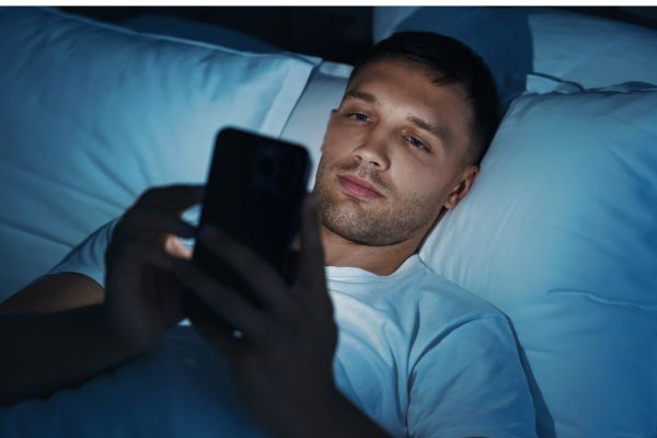 Young man lying in bed in the dark, using his cell phone with the blue light from the screen illuminating his face. The image illustrates bad habits of using smartphones before bed, highlighting the impacts on sleep quality.