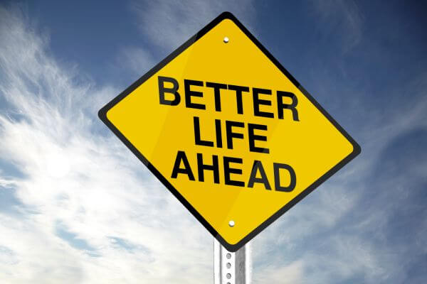 A yellow warning sign, shaped like a rotated square with one corner pointing downwards and a thick black border. The phrase 'BETTER LIFE AHEAD' is written in bold, black, uppercase letters, split into three lines. The sign is attached to a metal pole and is seen against a clear blue sky with a few sparse white clouds.
