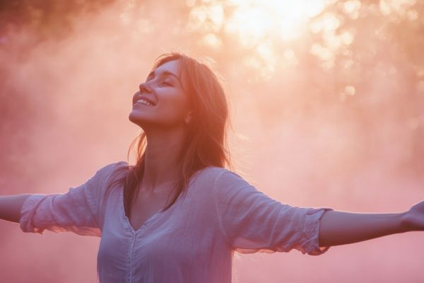 Young woman smiling and looking upwards with arms outstretched against a backdrop of golden/rosy light, conveying freedom, happiness, and a sense of purpose.