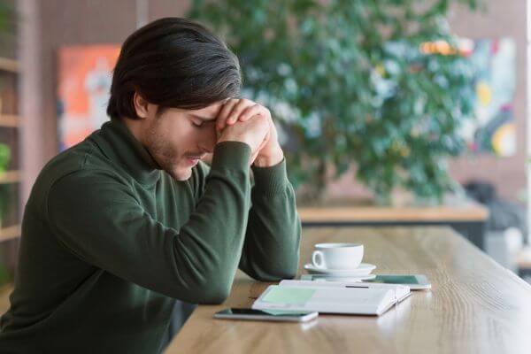A man with dark hair, wearing a green turtleneck, sitting at a wooden table with his head bowed and hands clasped over his forehead, appearing deeply troubled or contemplative. A white coffee cup, a notebook, and a smartphone are on the table. This image conveys feelings of stress or overwhelm, highlighting the challenging moments that call for inner resilience.